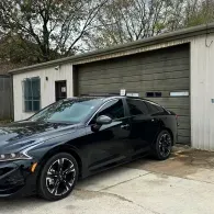 Black sedan parked in front of a garage with a closed door and wooded background