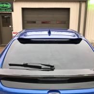 Blue car parked in front of a garage door, viewed from the rear with a rear spoiler and tinted window.