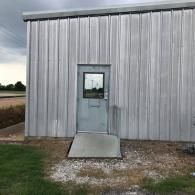 Metal building exterior with a small door and ramp, set beside gravel and grass