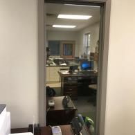 Office doorway looking into a cubicle workspace with desks, computer monitors, and fluorescent lights.