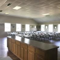 Empty banquet hall with rows of folding chairs and a wooden serving counter under fluorescent lights