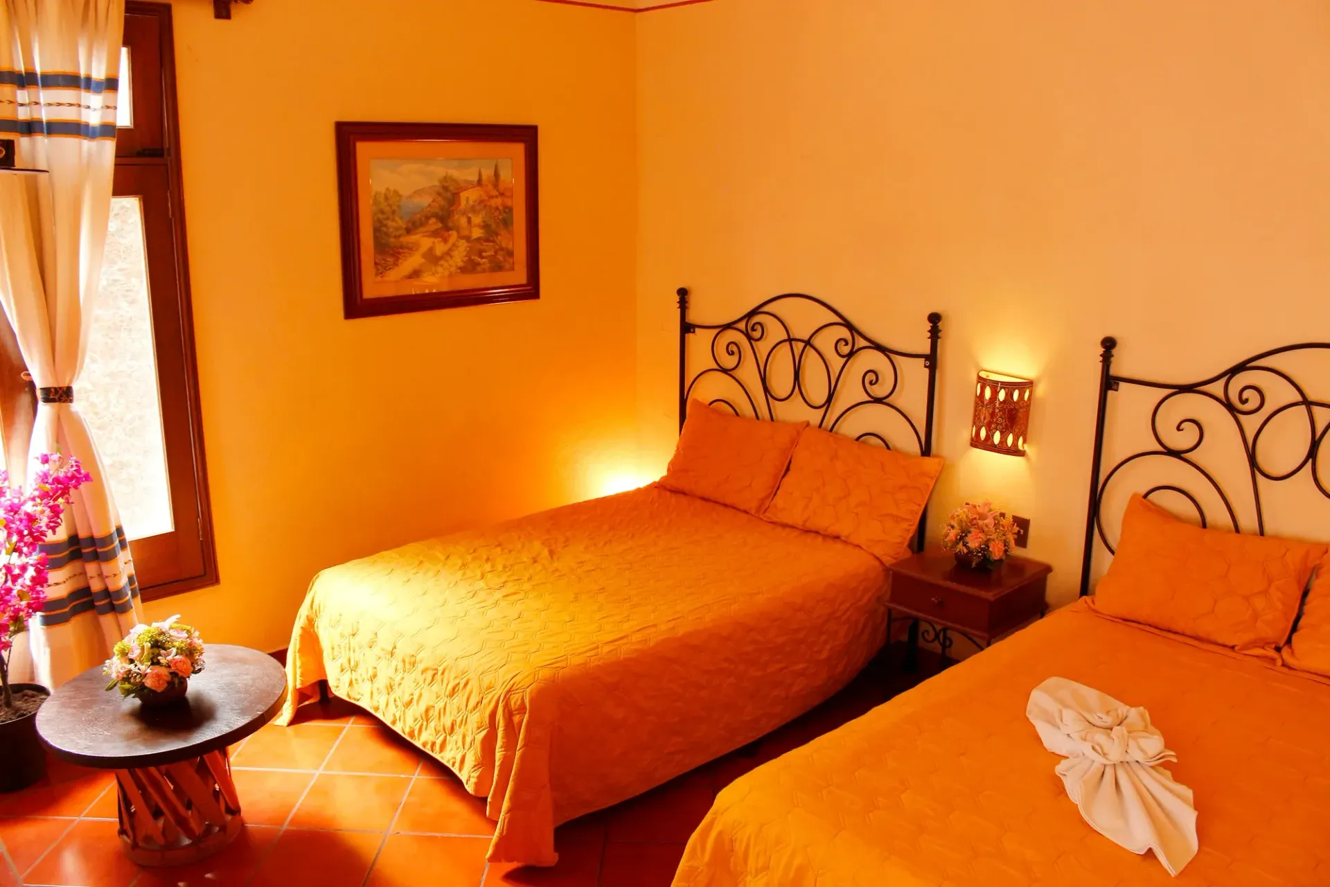 Un dormitorio con dos camas, ropa de cama de color naranja, cabeceros de hierro forjado, iluminación cálida.