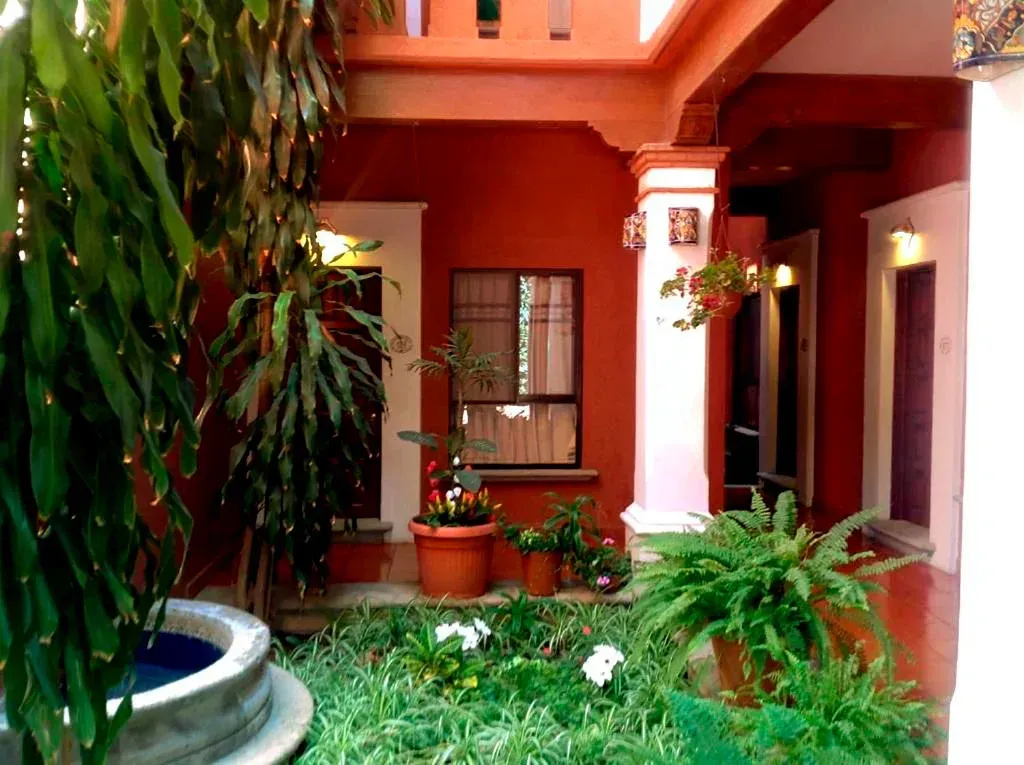 A courtyard with terracotta walls, a small fountain, lush green plants in pots, and white pillars under a covered walkway.