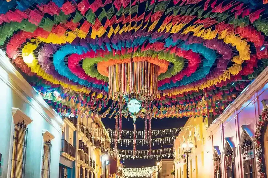 A street at night decorated with vibrant, circular hanging papel picado banners in rainbow colors.