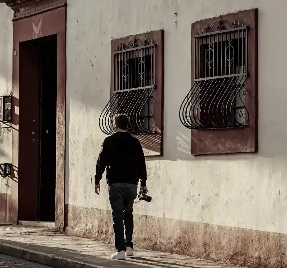 A person holding a camera walks along a beige building with two decorative, wrought-iron caged windows and a tall doorway.