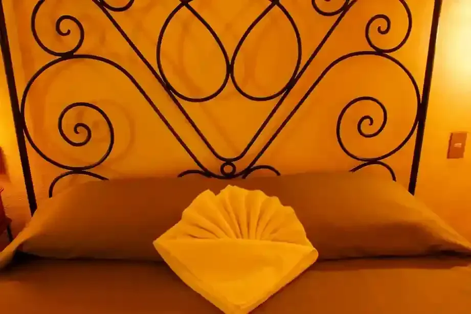 A bed with a gray sheet and a decorative, fan-folded white towel in front of a black metal scrolled headboard.