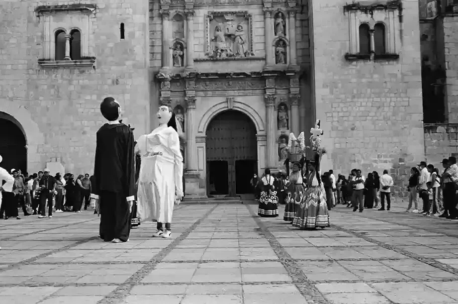 Two large, costumed figures stand in a stone plaza in front of an ornate historic church, watched by a crowd of onlookers.