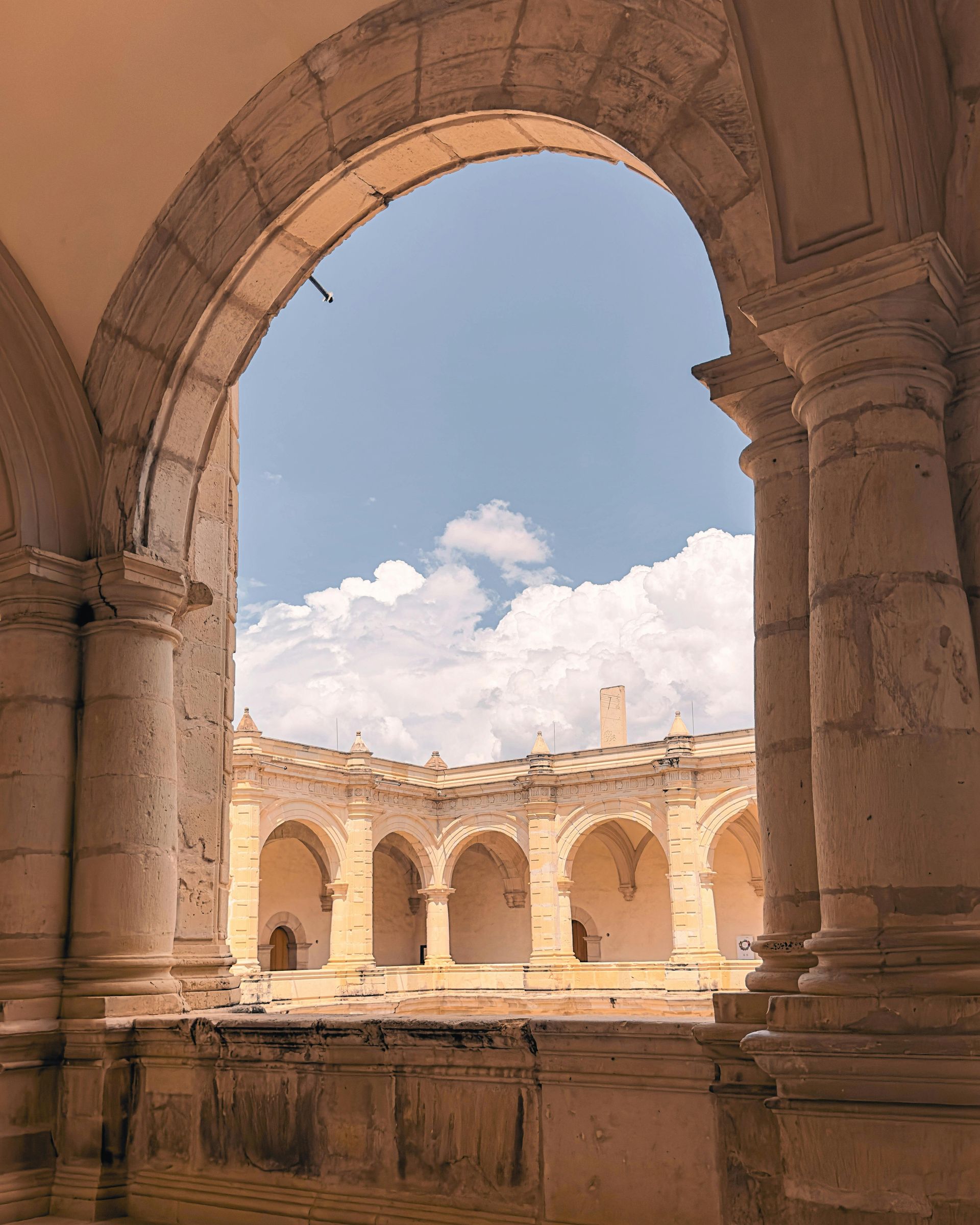 Vista de un antiguo patio de piedra beige con columnatas arqueadas bajo un cielo azul brillante con nubes blancas.