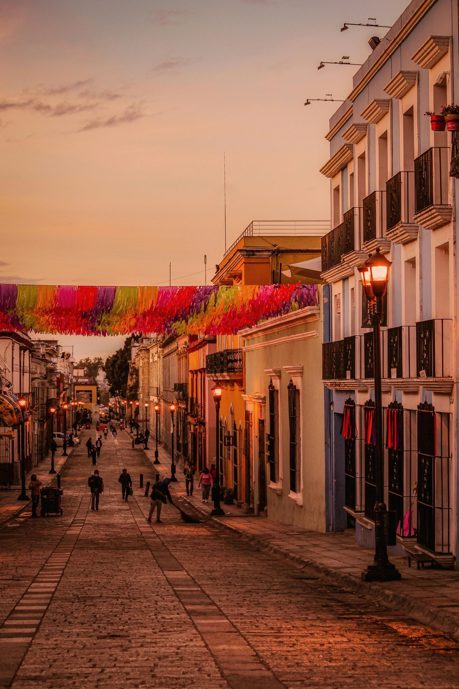 Una calle empedrada al atardecer, bordeada de edificios coloniales y adornada con coloridas guirnaldas de papel picado.