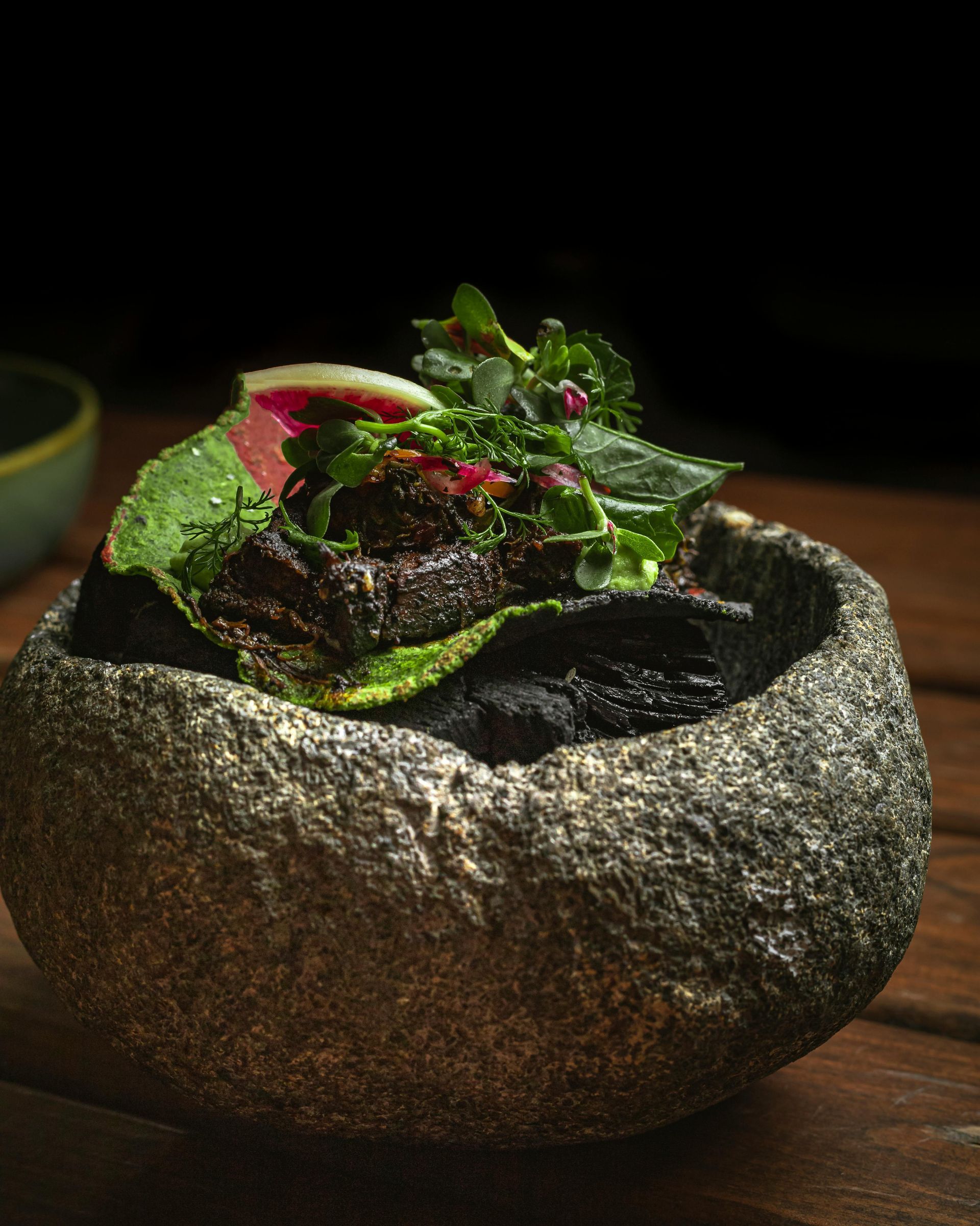 Un cuenco de piedra oscura y texturizada contiene carne sellada, cubierta con hierbas verdes vibrantes.