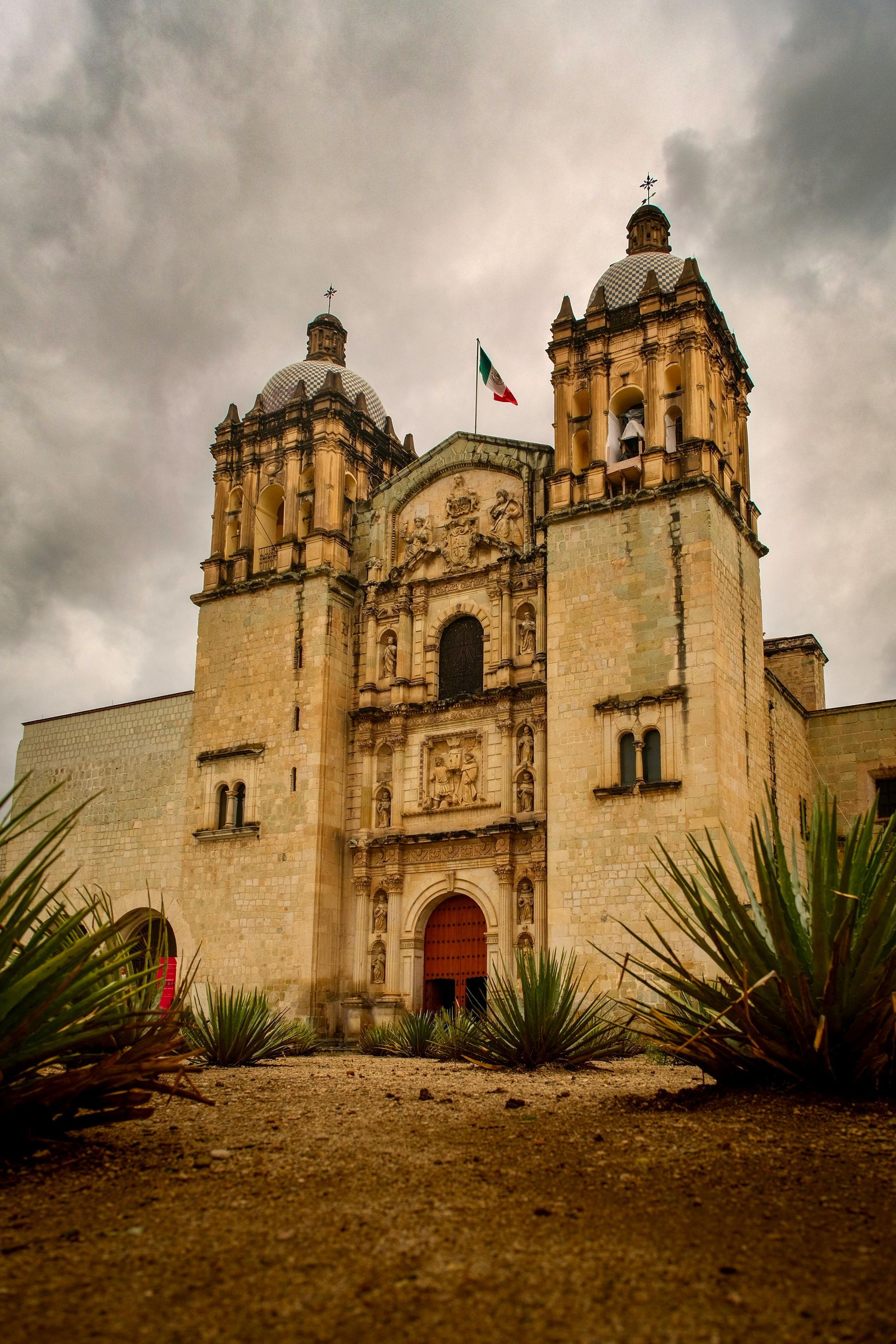 La histórica iglesia de piedra de Santo Domingo de Guzmán en Oaxaca, México, con sus dos torres bajo un cielo nublado.
