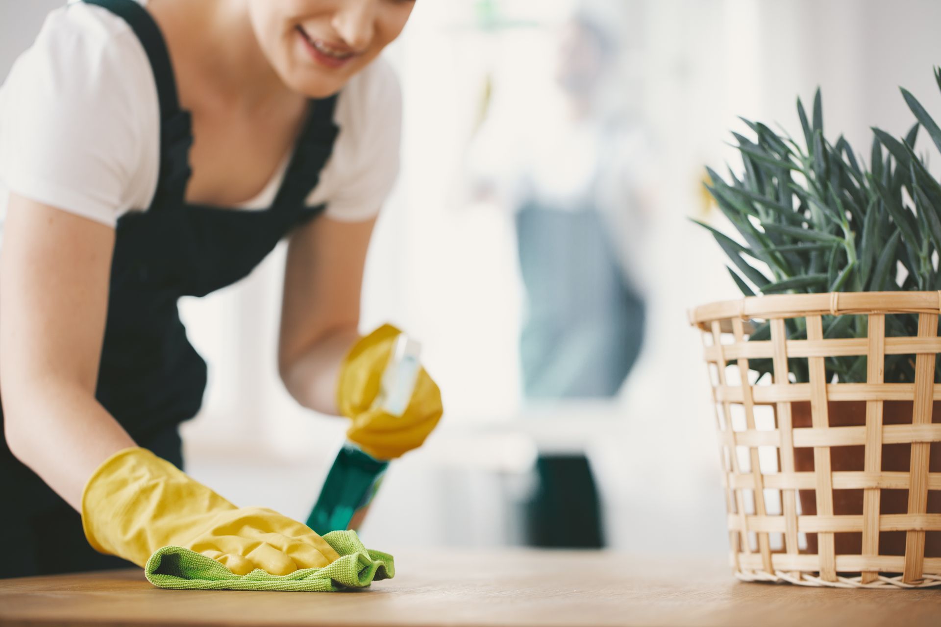 Woman cleaning a wooden table with spray bottle and cloth, smiling, wearing yellow gloves, potted plant in background.