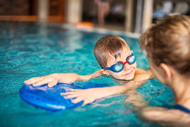 Child holding a blue kickboard in a swimming pool while another person watches nearby