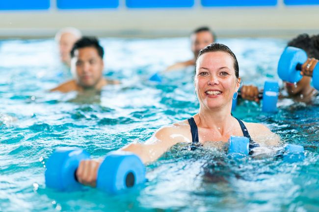 People doing water aerobics in a pool, holding blue foam weights, smiling at the camera
