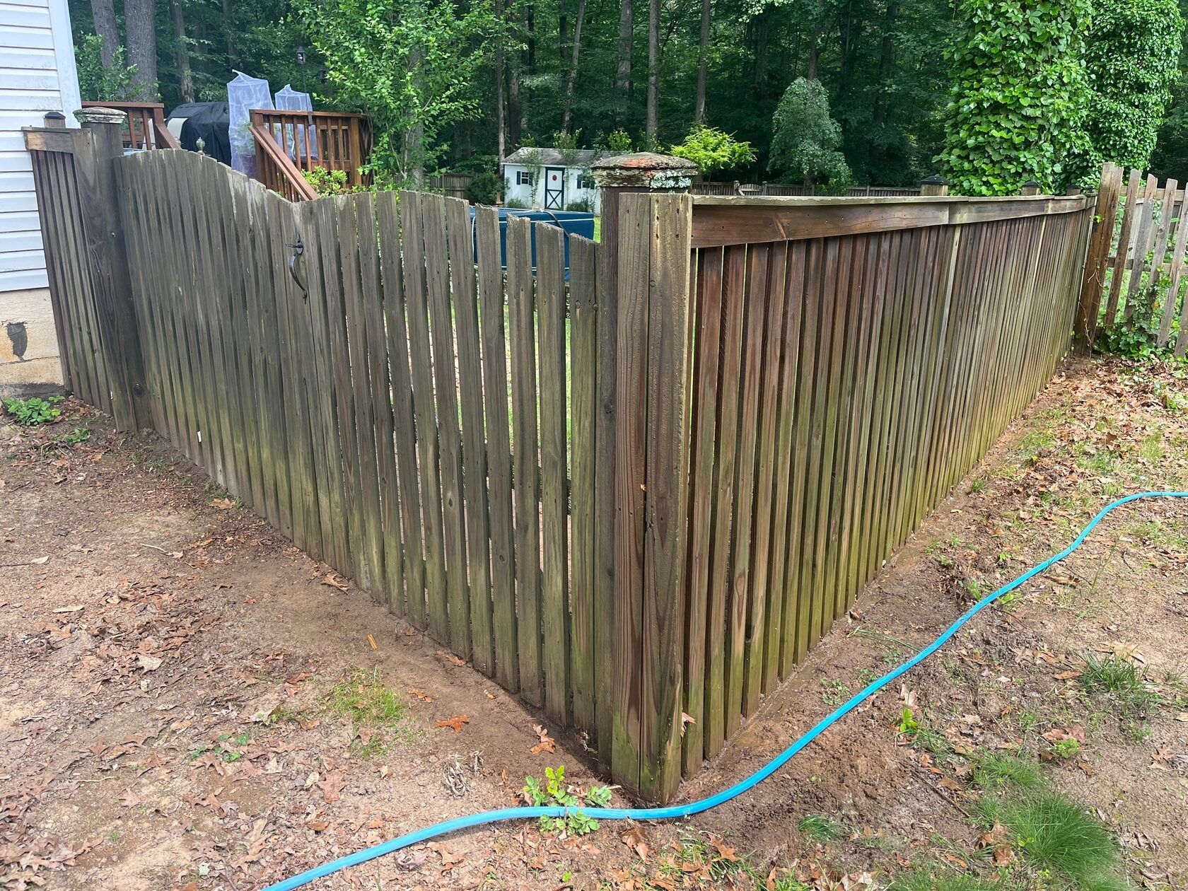 Wooden fence corner in a yard, weathered and shadowed, with a blue garden hose.