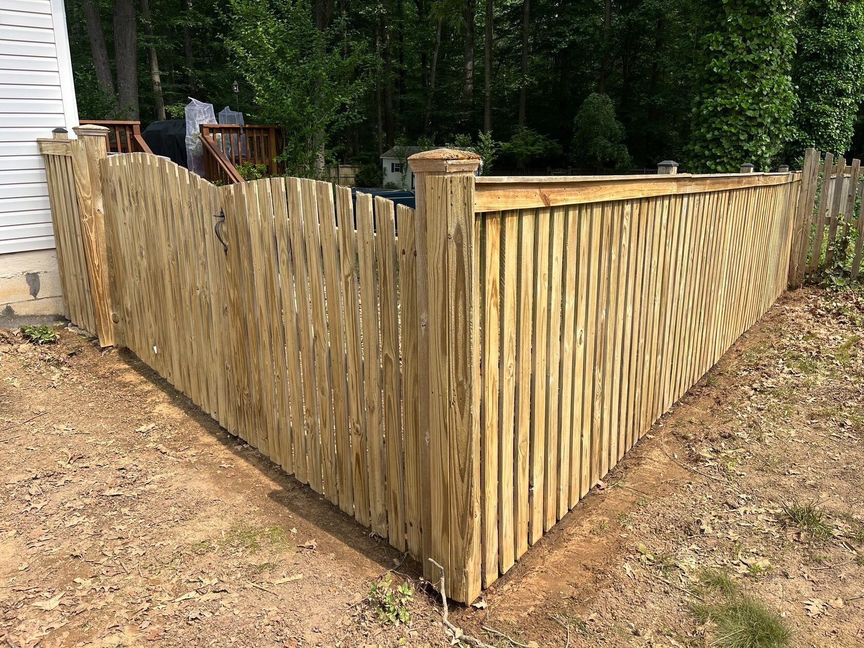 Wooden fence around a yard corner. Natural wood color. Green trees in the background, sunny outdoor setting.