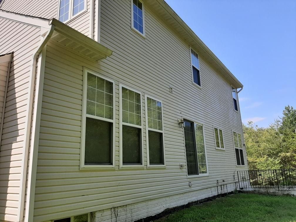 Two-story beige house with multiple windows on a sunny day. Green grass and trees are visible.