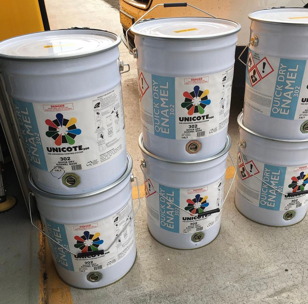 Stacked white chemical buckets on a floor, labeled Unicote Enamel, with a yellow lid in the background