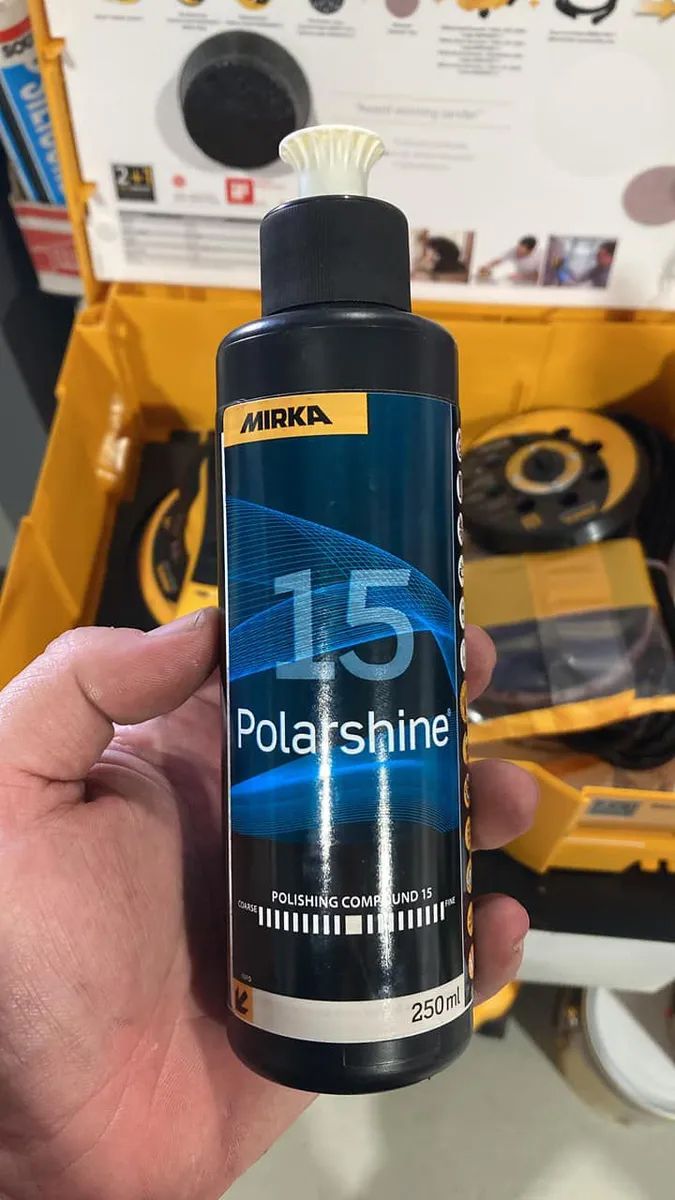 Hand Holding a Black Aeka 15 Polishine Bottle in Front of a Yellow Polishing Machine — Paint Place Cowra in Bathurst, NSW