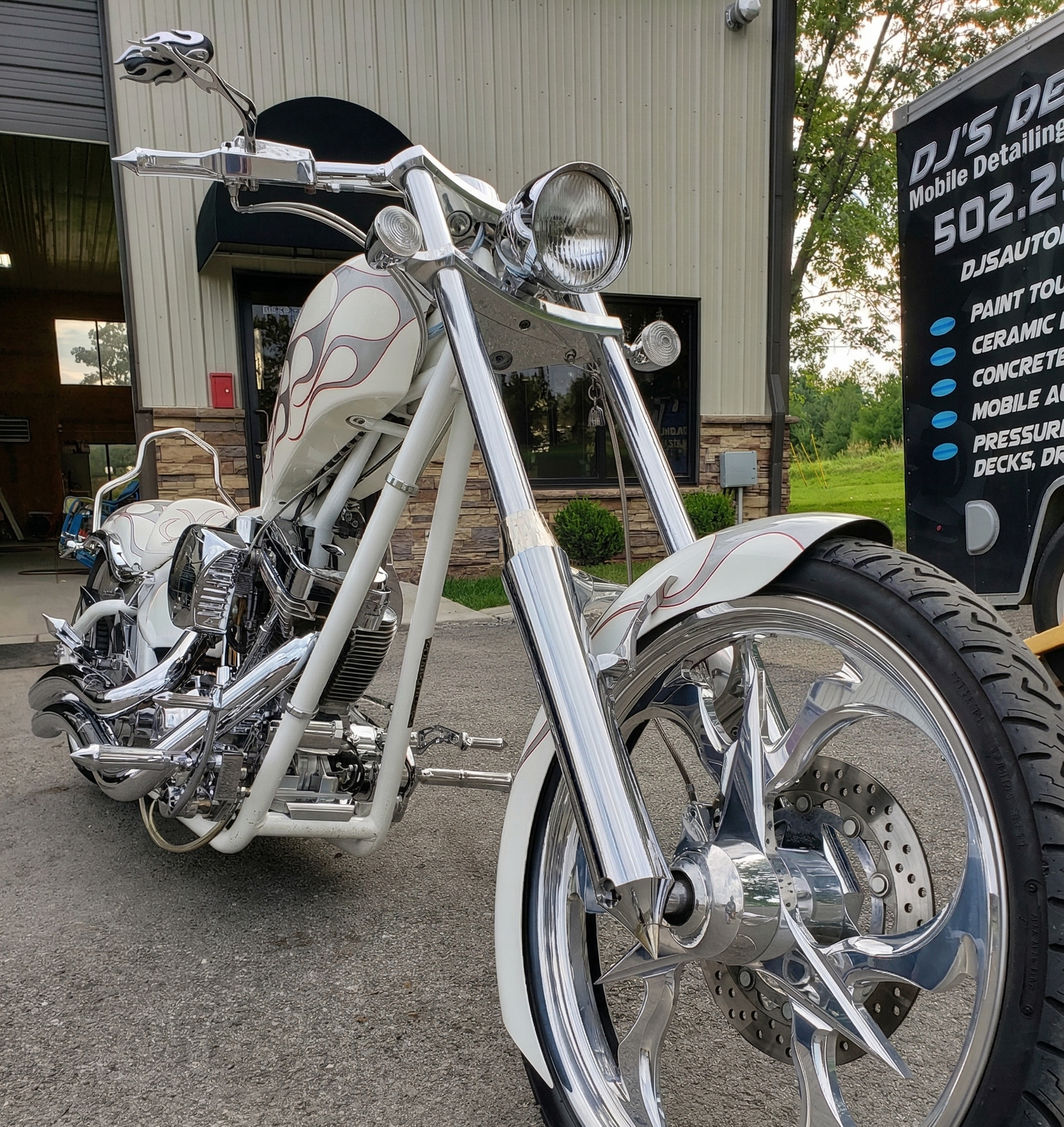 Custom white chopper motorcycle with chrome wheels and flame detailing, parked outside a detailing shop.