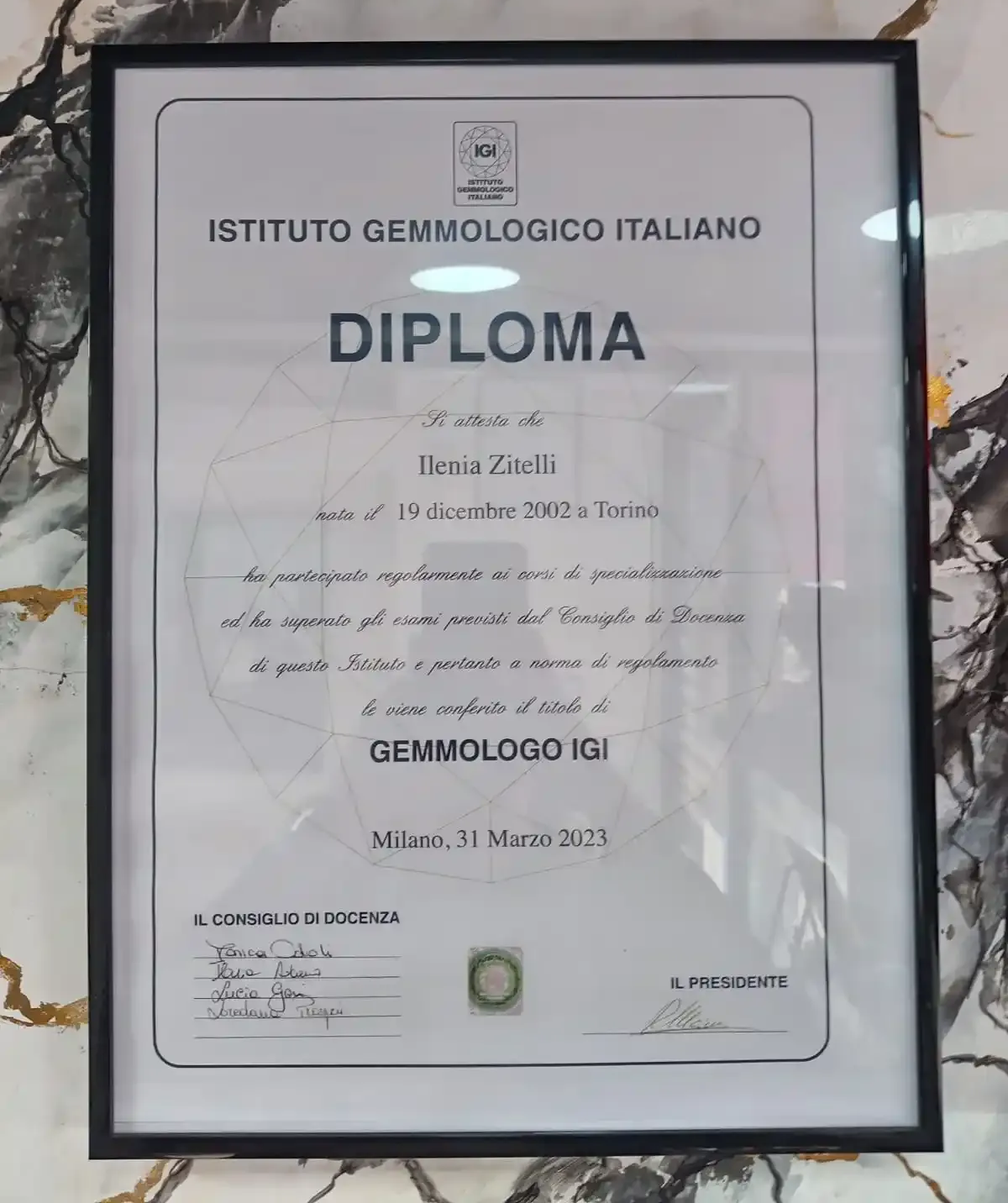 Diploma dell'Istituto Gemmologico incorniciato con certificato bianco e bordo nero, montato su una parete di marmo.