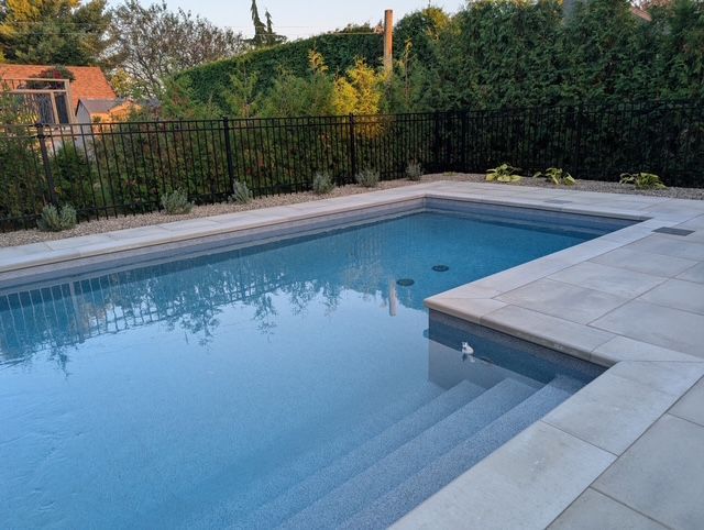 A rectangular pool with light blue water and steps, surrounded by stone and a black fence.
