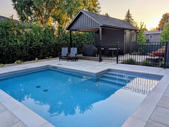 Backyard pool with black pergola, lounge chairs, and fencing, set against greenery and a house.