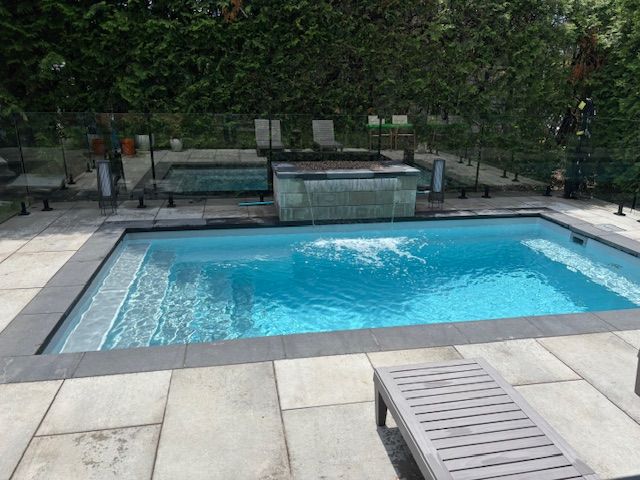 Rectangular blue pool with waterfall, stone patio, lounge chair, and glass fence.