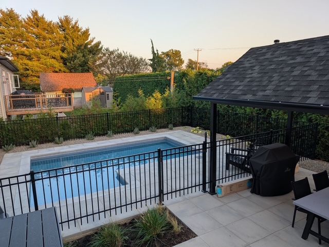 Backyard pool surrounded by black fence and patio, with a covered grill area, trees, and sky.