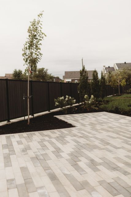 Patio with light-colored pavers, dark fence, small trees, and shrubs; cloudy sky.