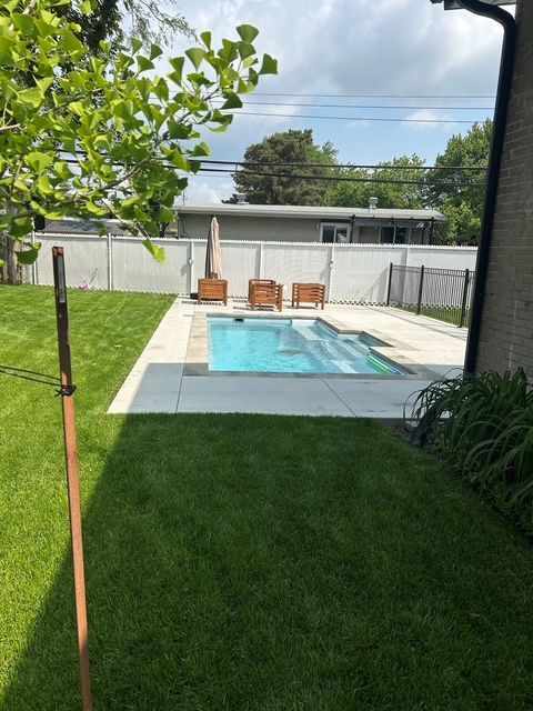 A rectangular swimming pool surrounded by a concrete patio in a backyard with green grass, wooden lounge chairs, and a white fence.