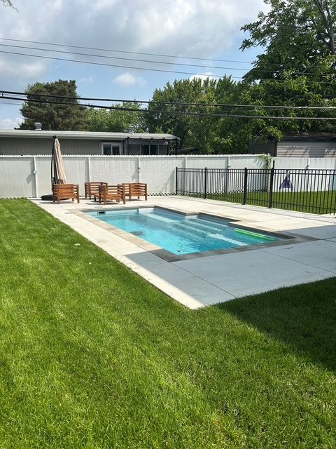 Backyard pool with light gray pavers, surrounded by green grass. Wooden furniture and black fence visible.