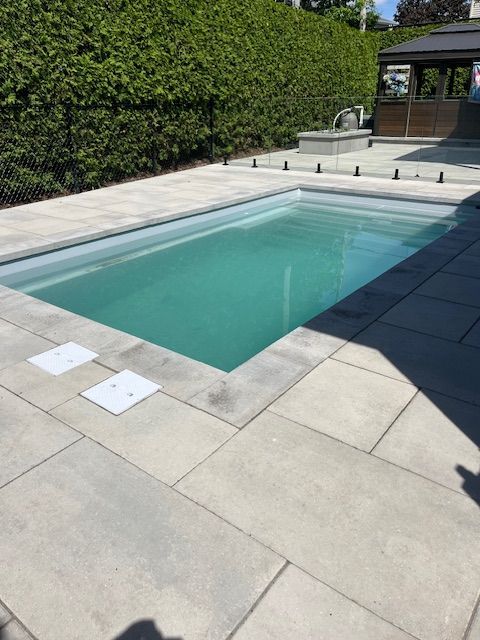 A rectangular backyard swimming pool surrounded by gray stone pavers. A green hedge is behind the pool.