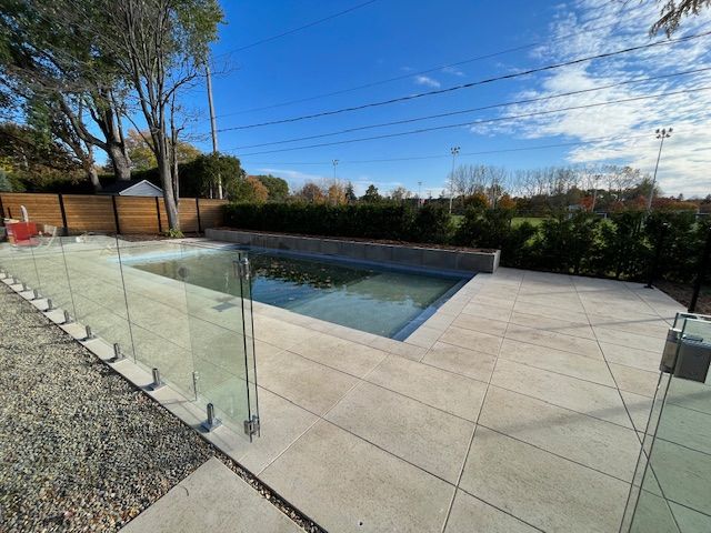 A rectangular swimming pool surrounded by a concrete patio and glass safety fence. Overcast sky.