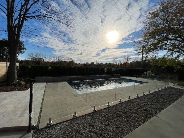 Pool surrounded by stone patio and glass fence under a sunny, cloudy sky.