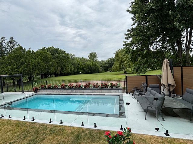 Rectangular pool with glass fence, patio furniture, and a grassy backyard under a cloudy sky.