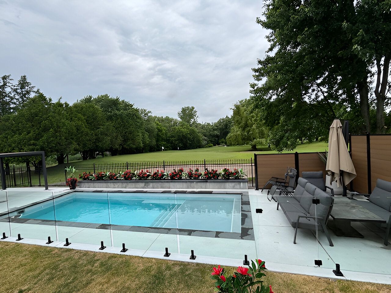 Pool with a glass fence, surrounded by a patio and lawn. In the background are trees and a cloudy sky.