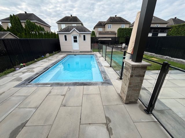 Rectangular backyard swimming pool surrounded by gray pavers and a black fence; small shed in background.