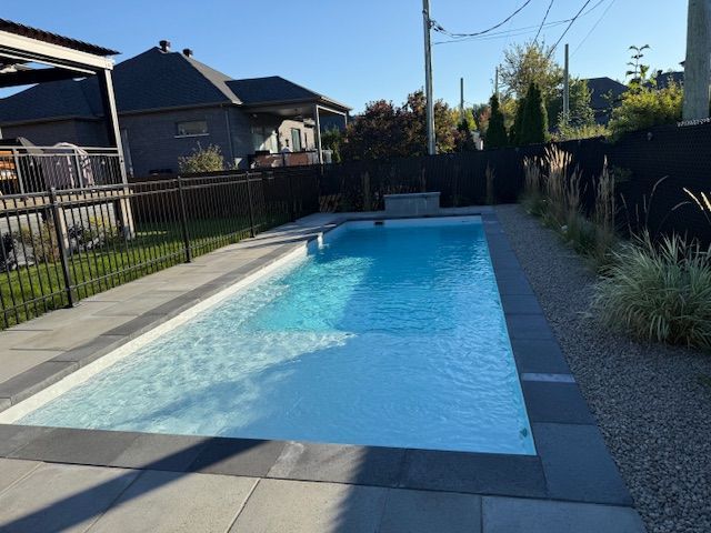 Rectangular in-ground pool surrounded by gray stone, with a black fence, gravel and a few plants in the background.