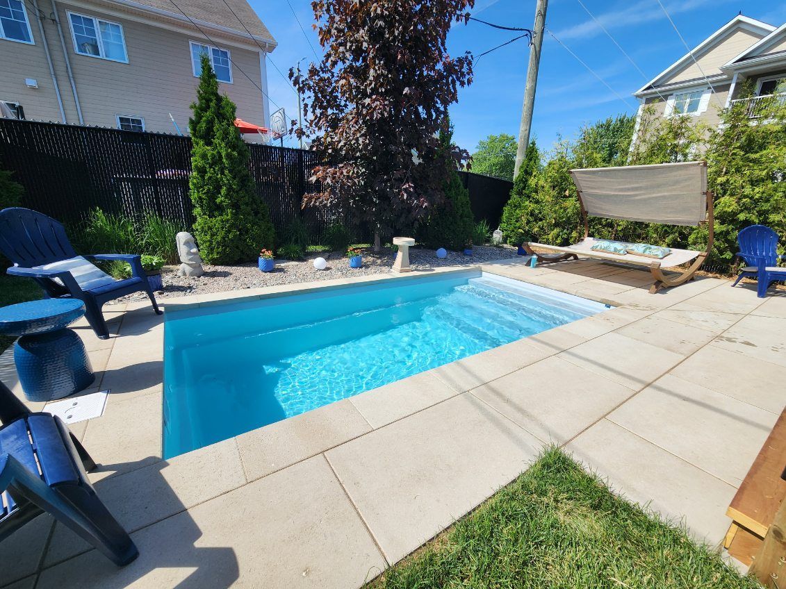 Small backyard pool with stairs, surrounded by a paved patio, chairs, and greenery.