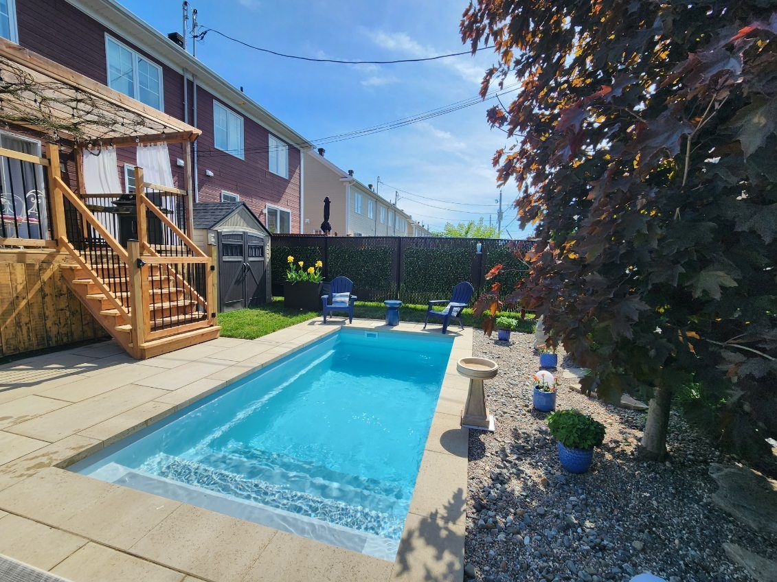 Backyard with a small pool, patio, and wooden deck under a sunny blue sky.