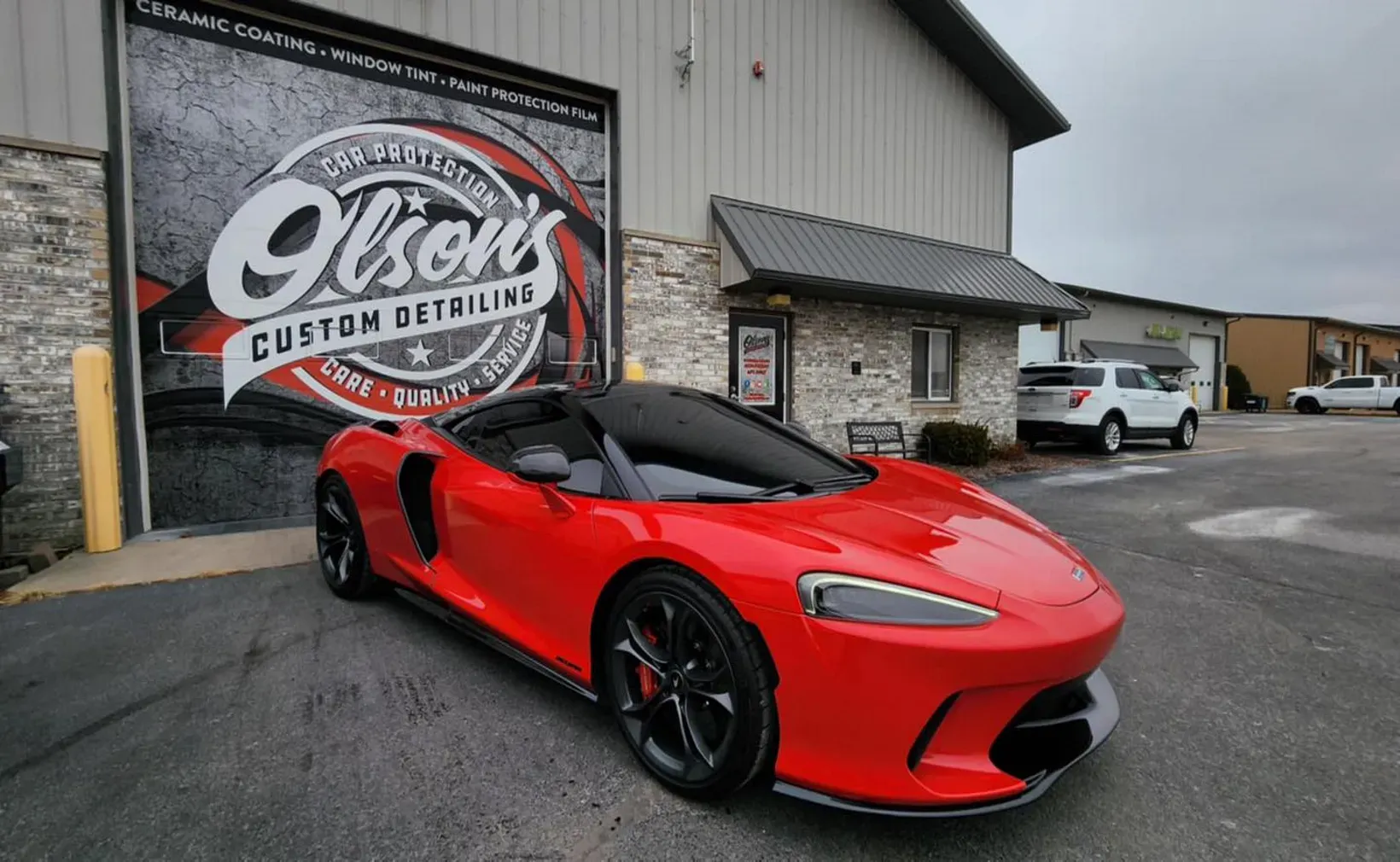Red McLaren sports car in front of a detailing shop with a white SUV in the background.