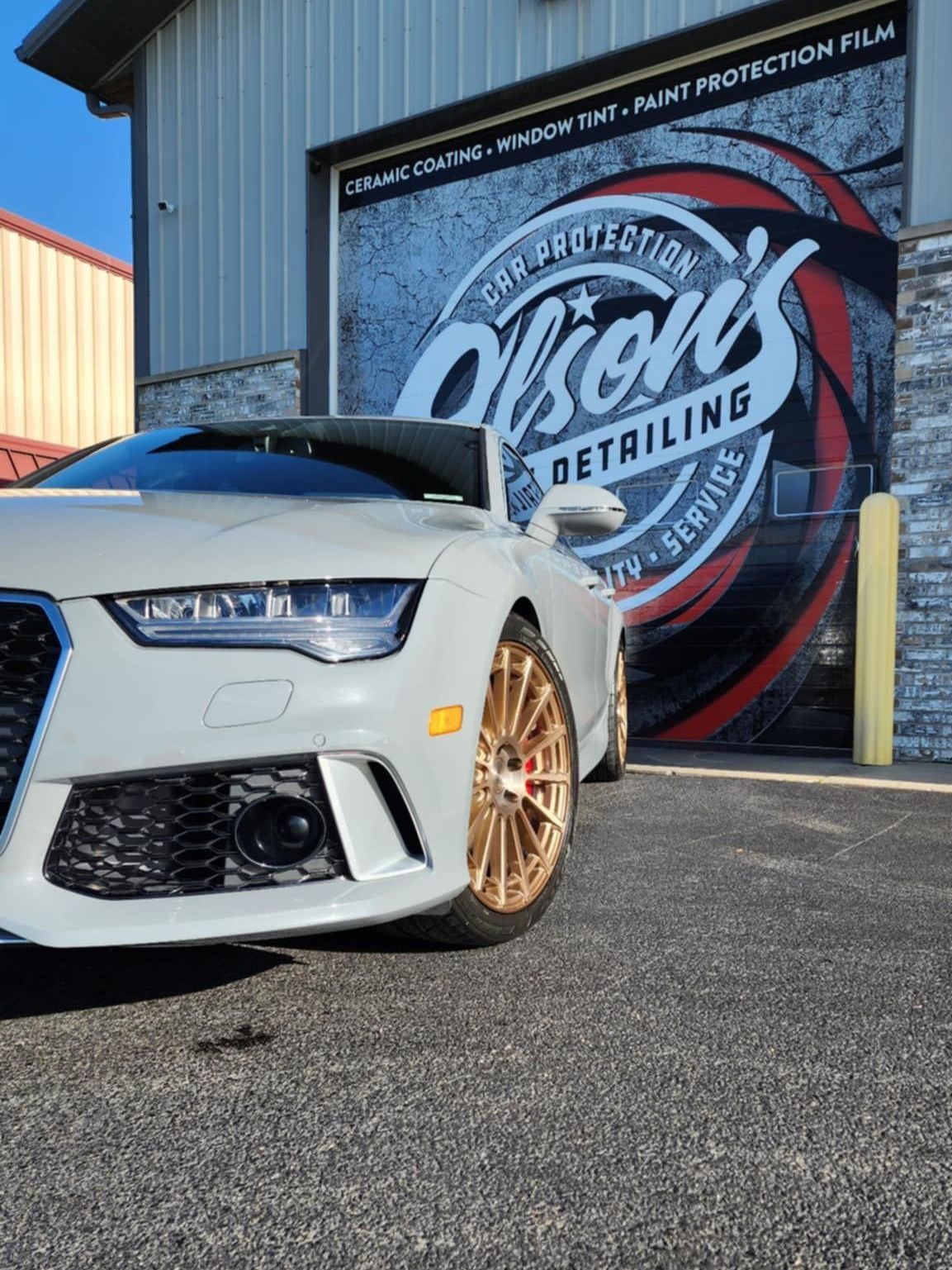 White Audi with gold rims parked in front of Olson's Detailing shop, featuring their logo.
