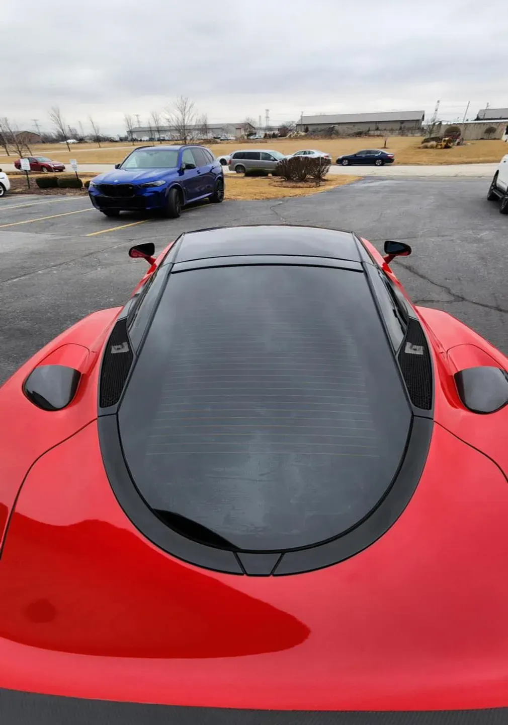 Red sports car with black roof, parked in a lot, other vehicles and buildings in the background.