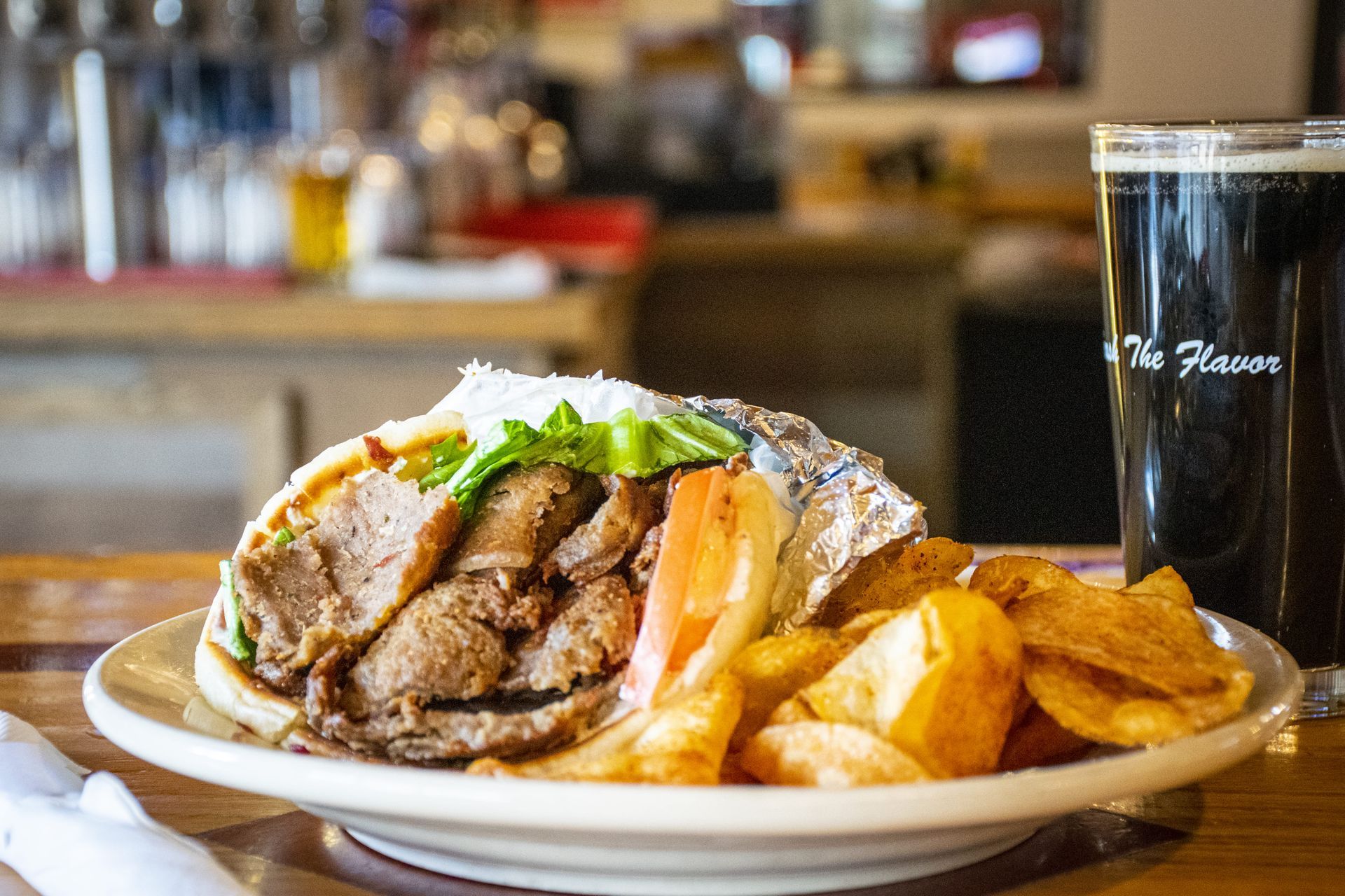 Plated sandwich with fries and dipping sauce beside a glass of dark beer on a bar counter