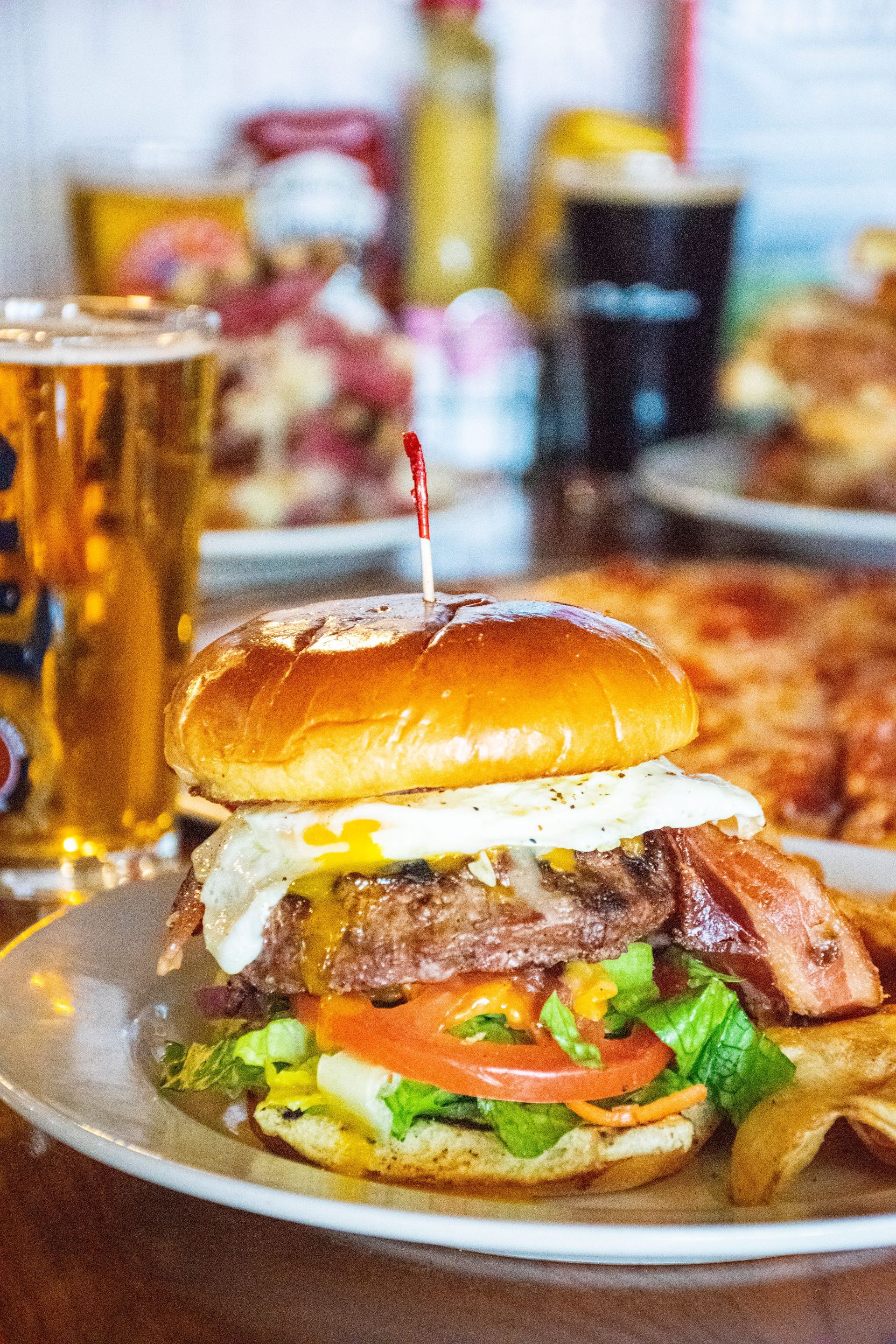 Burger with fried egg, bacon, lettuce, and tomato on a plate beside a glass of beer.