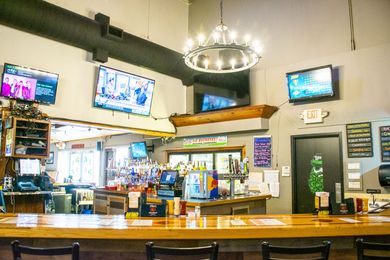 Empty restaurant bar with TV screens, wooden counter, hanging chandelier, and bright interior.
