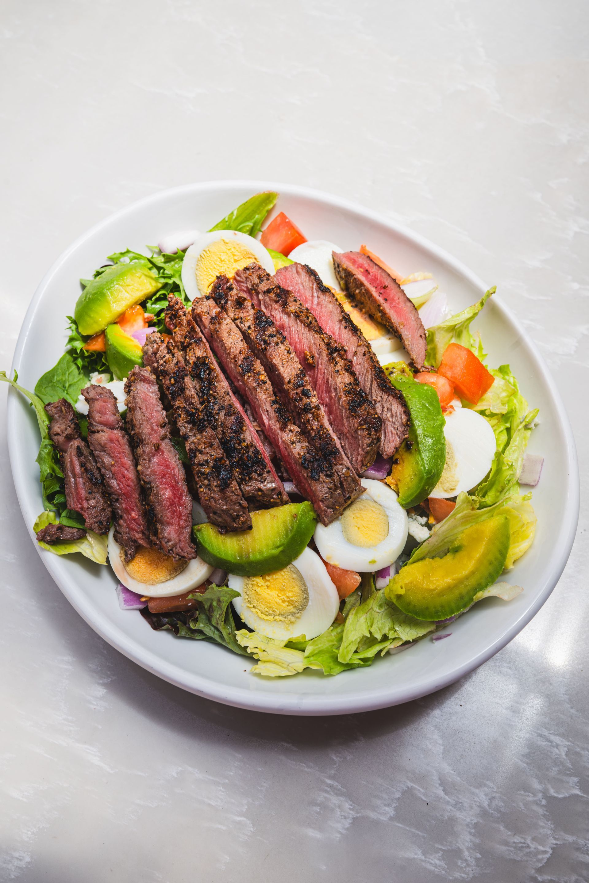 Plated salad with sliced grilled steak, hard-boiled eggs, avocado, and greens on a white plate