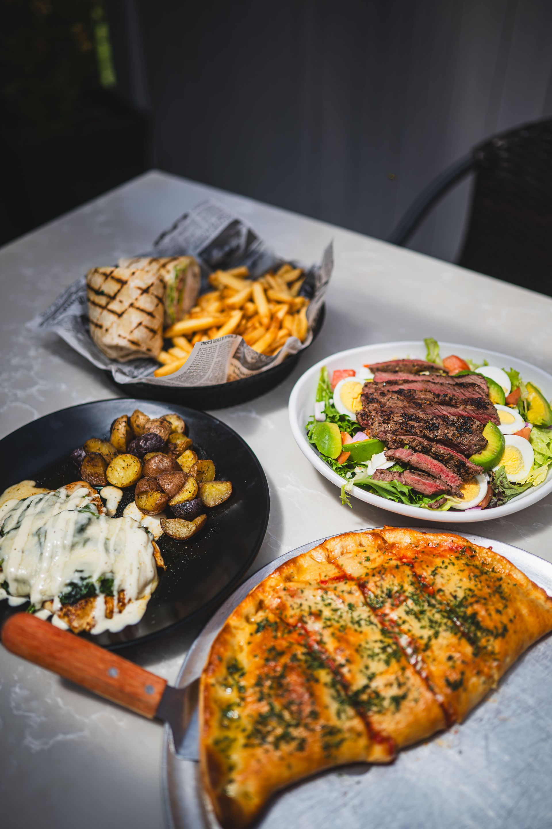 Assorted grilled dishes on a table: pizza, steak salad, fries, potatoes, and a cheese-topped skillet dish