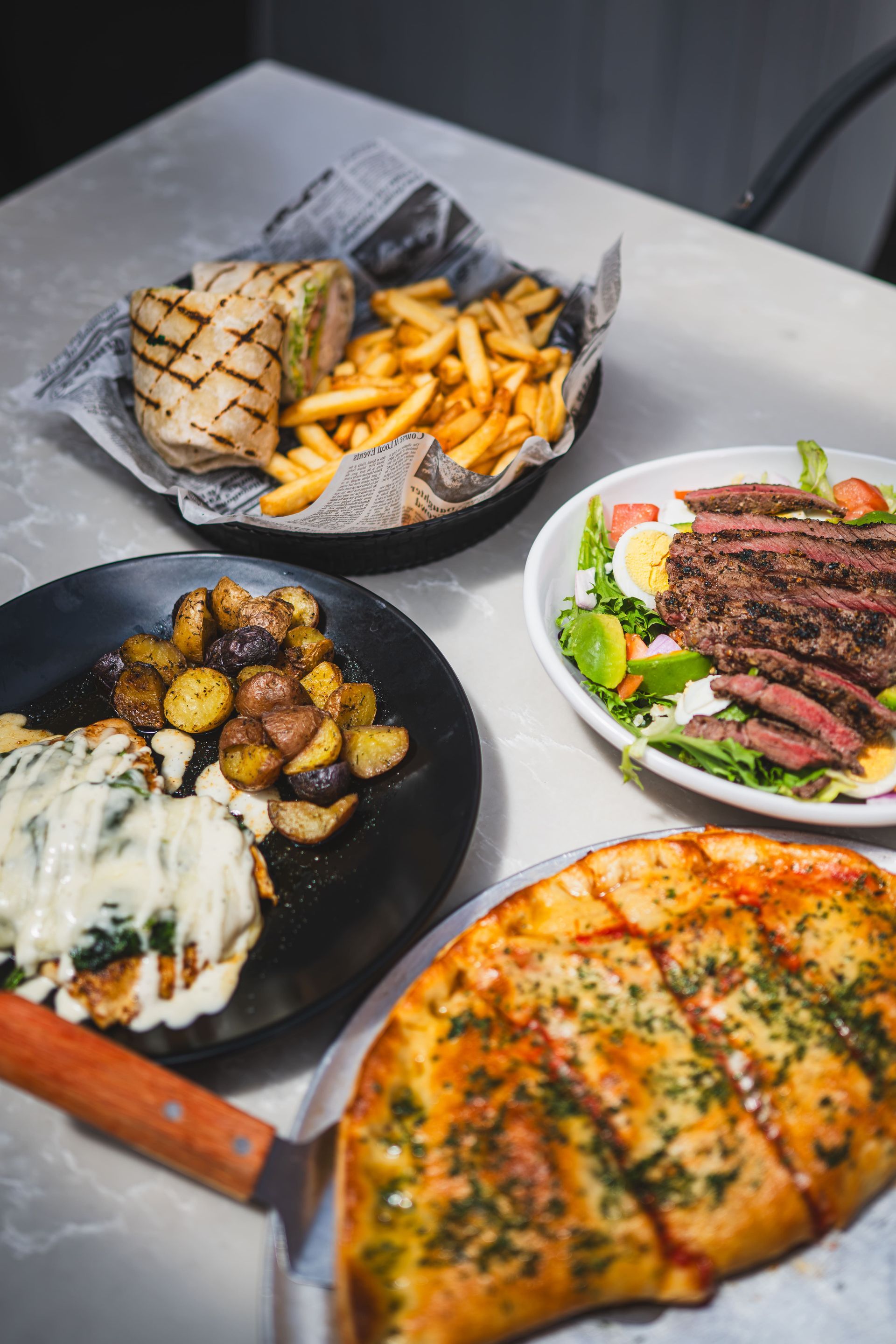 Assorted plated foods on a table: sandwich with fries, steak salad, roasted potatoes, and pizza halves
